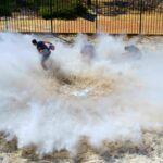 People running through a large dust/smoke cloud kicked up from the ground in an outdoor fenced area.