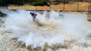 People running through a large dust/smoke cloud kicked up from the ground in an outdoor fenced area.