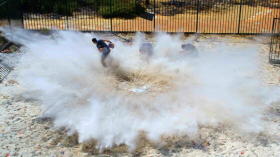 People running through a large dust/smoke cloud kicked up from the ground in an outdoor fenced area.