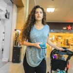A young woman with long curly hair pushes a baby stroller through a mall near a Macy's storefront, holding a blue smartphone in one hand.