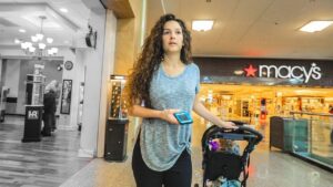 A young woman with long curly hair pushes a baby stroller through a mall near a Macy's storefront, holding a blue smartphone in one hand.