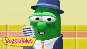 Green VeggieTales bean character wearing a blue hat and suit, smiling while singing into a vintage microphone on a colorful stage. Logo reads VeggieTales.