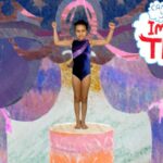 Young girl in a navy leotard stands on a pink pedestal with arms raised on a colorful stage backdrop, with the 'Imagine That' logo visible.