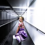 Young girl in a floral top with a purple jacket and small suitcase walks through a bright airport jet bridge, looking back over her shoulder.