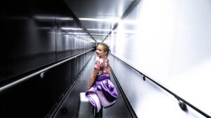 Young girl in a floral top with a purple jacket and small suitcase walks through a bright airport jet bridge, looking back over her shoulder.