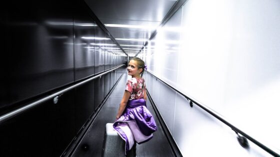 Young girl in a floral top with a purple jacket and small suitcase walks through a bright airport jet bridge, looking back over her shoulder.