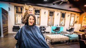 Woman seated in a salon cape while a stylist lifts her hair from behind, smiling at the camera in a modern hair salon setting.