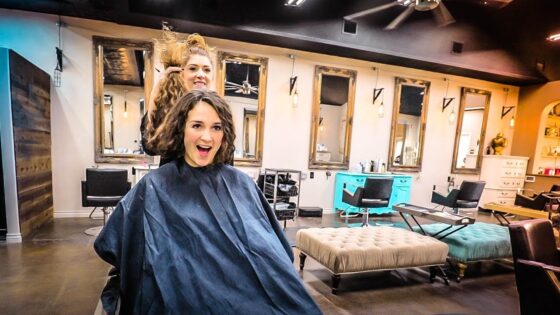 Woman seated in a salon cape while a stylist lifts her hair from behind, smiling at the camera in a modern hair salon setting.