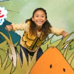 Girl in a yellow vest smiles as she leans through cartoon grass in a bright, illustrated landscape with the 'Imagine That!' logo on the left.