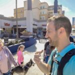 Man in teal shirt with backpack taking a selfie with a smiling group outdoors in a sunny shopping district, a dog nearby.