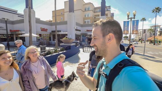Man in teal shirt with backpack taking a selfie with a smiling group outdoors in a sunny shopping district, a dog nearby.