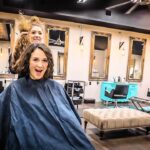 Client wearing a dark cape smiles at the camera while a hairstylist lifts a strand of hair from behind her in a modern salon with mirrors and seating in the background.