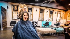 Client wearing a dark cape smiles at the camera while a hairstylist lifts a strand of hair from behind her in a modern salon with mirrors and seating in the background.