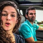 Two people in a car: a woman with curly hair in the passenger seat speaking to the camera while a man in a teal shirt drives.