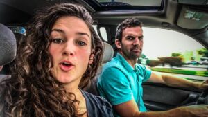 Two people in a car: a woman with curly hair in the passenger seat speaking to the camera while a man in a teal shirt drives.