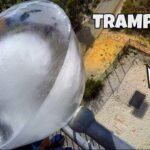 Aerial view of a tall transparent amusement-ride tube with a fenced area below and the word TRAMPOLINE at the top right.