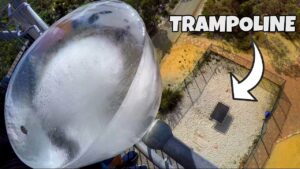 Aerial view of a tall transparent amusement-ride tube with a fenced area below and the word TRAMPOLINE at the top right.