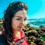 Woman with dark hair in a red jacket and backpack standing on rocky coastline by blue ocean.