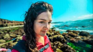 Woman with dark hair in a red jacket and backpack standing on rocky coastline by blue ocean.