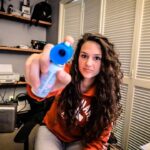 Woman with curly hair in an orange hoodie holds a blue-capped bottle toward the camera, likely eye drops or contact solution, in a home office setting.