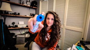 Woman with curly hair in an orange hoodie holds a blue-capped bottle toward the camera, likely eye drops or contact solution, in a home office setting.