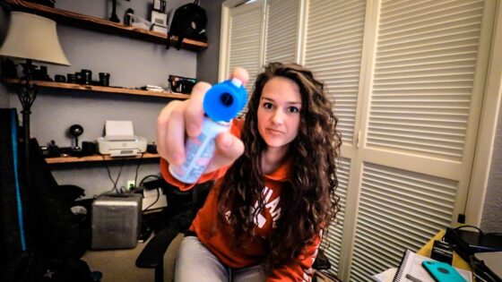 Woman with curly hair in an orange hoodie holds a blue-capped bottle toward the camera, likely eye drops or contact solution, in a home office setting.