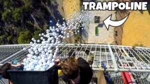 People on a tall metal platform dump a crate full of white balloons outward, creating a balloon spill below. A sign or graphic reading 'TRAMPOLINE' is visible in the corner.