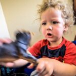 Toddler in a red shirt plays with a dark plastic dinosaur indoors, hands blurred from motion.