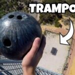 Person grips a dark bowling ball while leaning over a high railing, looking down at a courtyard below with an arrow overlay saying TRAMPOLINE.