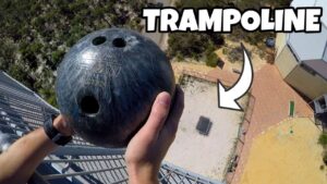 Person grips a dark bowling ball while leaning over a high railing, looking down at a courtyard below with an arrow overlay saying TRAMPOLINE.