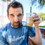 Man in a blue Billabong shirt holds an iced coffee with a straw, looking at the camera on an outdoor patio.