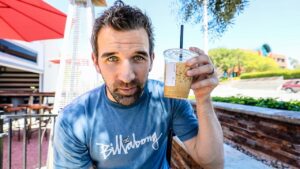 Man in a blue Billabong shirt holds an iced coffee with a straw, looking at the camera on an outdoor patio.