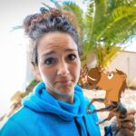 Young woman in a bright blue hoodie outdoors holding a small cartoon horse figure near her face, with palm trees in the background.