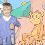 A smiling child in purple scrubs with pockets stands beside a cartoon monkey on a pink wall, with the 'Imagine That!' logo in a cloud on the left.