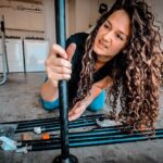 Woman kneels on a garage floor and assembles black metal poles for a DIY project, with tools and packaging nearby.
