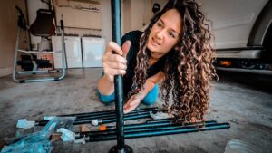 Woman kneels on a garage floor and assembles black metal poles for a DIY project, with tools and packaging nearby.