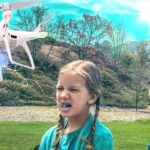 White quadcopter drone hovers near a girl with braids as she shouts, with a man in a green shirt controlling it in a park.