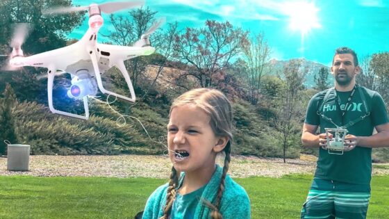 White quadcopter drone hovers near a girl with braids as she shouts, with a man in a green shirt controlling it in a park.
