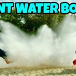 People outdoors get soaked by a huge water explosion; one person is mid‑air from the splash while two others kneel nearby with a water container. Bold text reads 'GIANT WATER BOMB!'