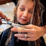 Hairdresser cutting a young girl's hair while the girl looks astonished, eyes wide as scissors hover near her strands