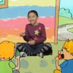 Center girl storyteller: a smiling young girl in a maroon star-pattern sweater sits cross-legged, surrounded by four kids eating apples in a bright, hand-drawn backdrop.