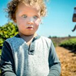 Young child with curly hair and blue eyes stands in a sunlit field, with a man holding a baby in the background.