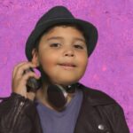 Young boy wearing a black hat and headphones around his neck, posing against a purple textured background.