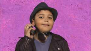 Young boy wearing a black hat and headphones around his neck, posing against a purple textured background.