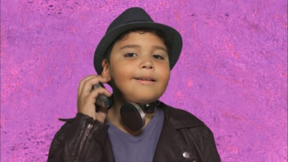 Young boy wearing a black hat and headphones around his neck, posing against a purple textured background.