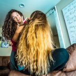 Woman with curly hair cuts another person’s long blonde hair in a casual living room scene, sitting on a brown couch by family photos and artwork.