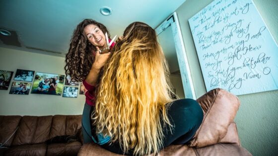 Woman with curly hair cuts another person’s long blonde hair in a casual living room scene, sitting on a brown couch by family photos and artwork.