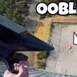 Person on a tall structure holds a large black anchor-like tool over the edge, with a fenced gravel area and a pink-green ball below; a bold 'OBLECK' thumbnail label is visible ahead of the scene.