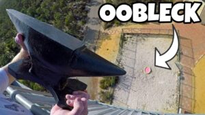 Person on a tall structure holds a large black anchor-like tool over the edge, with a fenced gravel area and a pink-green ball below; a bold 'OBLECK' thumbnail label is visible ahead of the scene.
