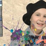 Smiling girl in a black beret with multicolor paint splatters on her shirt, in front of a cartoon window background.